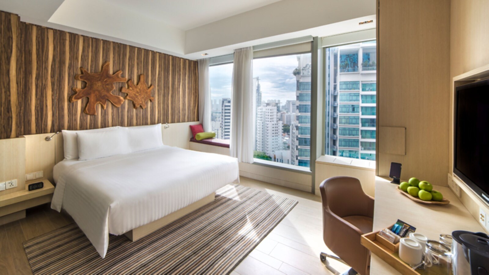 A hotel room with a large bed, a wooden accent wall, a city view, a desk with a chair, and a television. - Oasia Hotel Novena, Singapore (Singapore)
